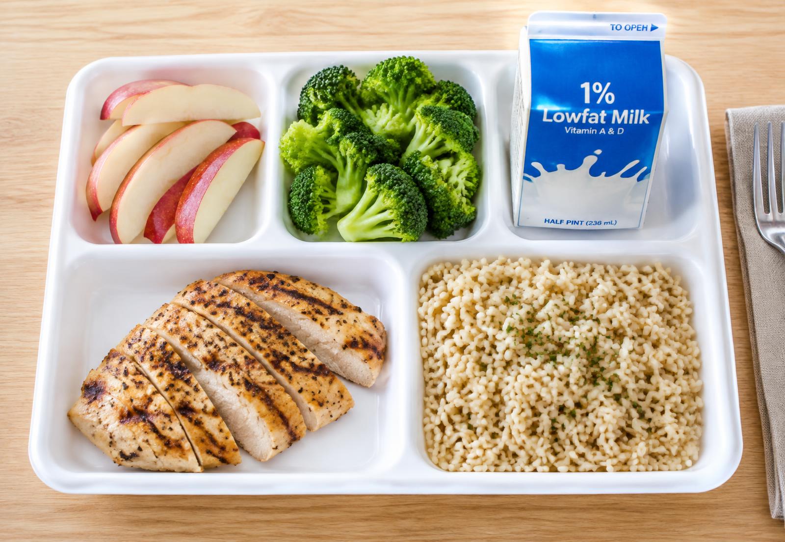 A school lunch tray with grilled chicken, rice, broccoli, apple slices and milk